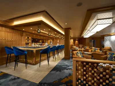 Die Maltings Whiskey Bar an Bord der Norwegian Bliss. Blaue Samt-Barhocker am Tresen. Warme Holzwände, indirekte Beleuchtung und Sitzecken mit ockerfarbenen Sesseln prägen den eleganten Raum.