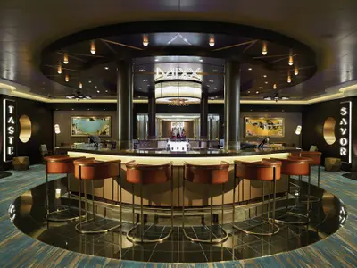 Die elegante Mixx Bar an Bord der Norwegian Bliss zeigt eine zentrale, runde Theke mit Barhockern, umgeben von dunklen Holzelementen, Kunstwerken und markanter Beleuchtung.