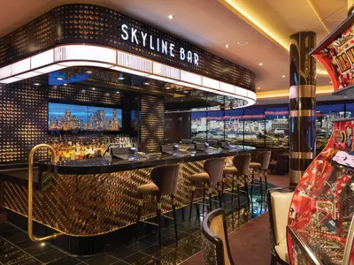 Die Skyline Bar auf der Norwegian Bliss zeigt eine moderne Bar mit Goldakzenten, Flaschen, Barhockern, Blick auf eine Skyline und rote Spielautomaten.