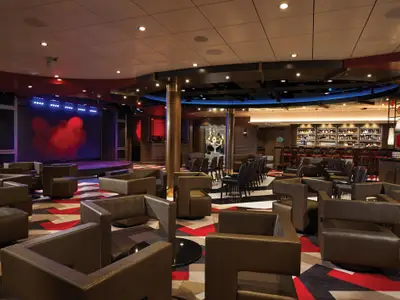 Die moderne Social Lounge an Bord der Norwegian Bliss mit beleuchteter Bühne und bequemen Lounge-Sesseln. Eine voll ausgestattete Bar ist im Hintergrund zu sehen. Das Ambiente ist einladend.