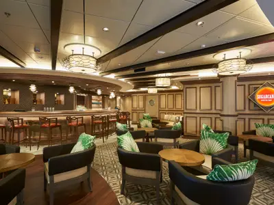 Innenansicht der Sugarcane Mojito Bar an Bord der Norwegian Bliss. Ein eleganter Raum mit einer langen Bar, Barhockern und Sitzbereichen mit Tischen, Sesseln und grünen Blattkissen.