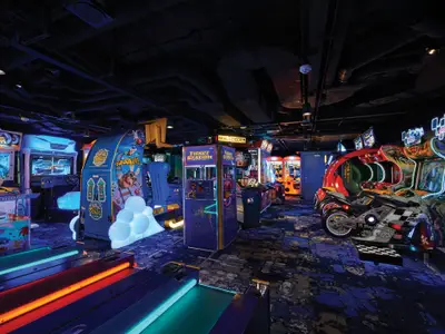 Ein breiter Blick in die beleuchtete Videospielhalle an Bord der Norwegian Bliss, gefüllt mit Rennspielen, Schiessbuden und Ticket-Automaten. Die Spielgeräte leuchten in Neonfarben und spiegeln sich auf dem gemusterten Teppichboden. An der Decke sind Lüftungsrohre und Beleuchtung sichtbar, was der Szene eine moderne, industrielle Ästhetik verleiht.
