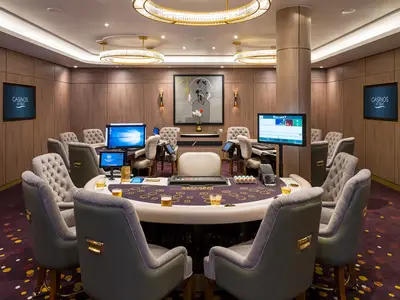 Baccarat-Casino an Bord der Norwegian Bliss. Runder Spieltisch, Monitore, Chips und Getraenke. Gepolsterte Stuehle. Holzwaende mit Bildschirmen, Kunstwerk. Eleganter Innenraum.