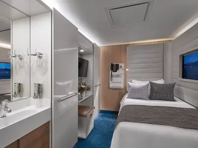 Helle, moderne Einzelkabine an Bord der Norwegian Bliss mit einem Einzelbett und gepolstertem Kopfteil. Links Waschbecken und Spiegel, daneben ein Fernseher. Ein Fenster bietet Ausblick auf eine abendliche Landschaft.