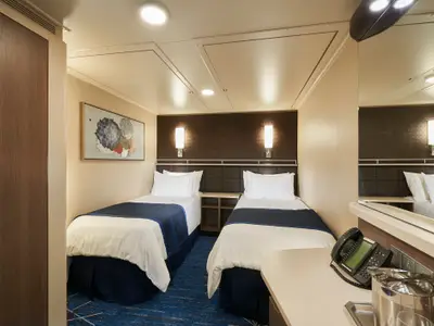 Gemuetliche Innenkabine an Bord der Norwegian Bliss mit zwei Einzelbetten, Nachttisch, Telefon und Wandkunst. Modernes Design mit blauen Akzenten und warmer Beleuchtung.