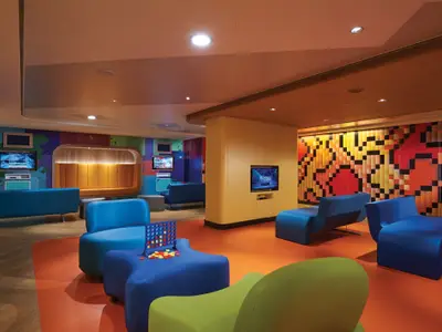 Ein bunter, moderner Kinderbereich namens Splash Academy an Bord der Norwegian Bliss. Der Raum ist mit weichen, organisch geformten Sitzelementen in Blau und Grün ausgestattet. Eine Connect Four-Spielkonsole steht auf einem blauen Hocker. An den Wänden sind Bildschirme mit Videospielen zu sehen, darunter Rocket League. Die Wände sind mit abstrakten, farbenfrohen Mustern im Pixel-Stil und Farbspritzern gestaltet. Die Decke ist hell und mit Einbauleuchten versehen, der Boden ist teils orange, teils holzfarben. Es sind keine Personen zu sehen.
