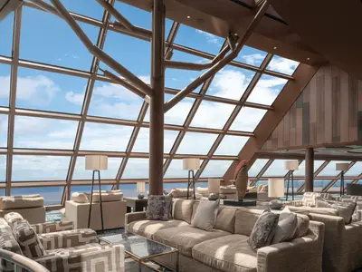Eine helle, geräumige Lounge an Bord der Norwegian Bliss mit beigefarbenen Sofas und Sesseln. Große Glasfenster bieten einen weiten Blick auf das blaue Meer und den Himmel. Die Holzstruktur im Deckenbereich erinnert an Äste.