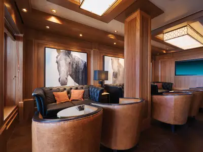 Stilvolle Lounge an Bord der Norwegian Bliss. Warme Holzvertäfelung, dunkle Ledersofas und braune Sessel. Grosse Pferdeporträts und ein Bildschirm schmücken den Raum.