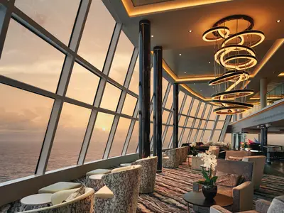 Geräumige Observation Lounge der Norwegian Bliss bei Sonnenuntergang über dem Meer. Grosse Fensterfront, elegante Sessel und spiralförmige Lichtinstallation an der Decke.