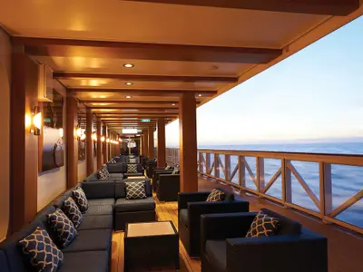 Ueberdachte Aussenlounge der Norwegian Bliss in der Abenddaemmerung. Reihen blauer Sitzgruppen mit Kissen unter beleuchteter Holzdecke bieten weiten Meerblick.