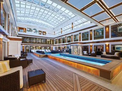 Geräumiger, luxuriöser Poolbereich unter einem Glasdach an Bord der Norwegian Bliss. Mit eleganten Holzdetails, bequemen Lounge-Möbeln und warmen Lichtakzenten.