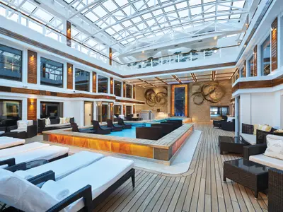Exklusiver Innenpoolbereich im Haven Courtyard an Bord der Norwegian Bliss. Mit Glasdach, beleuchtetem Pool, bequemen Liegen und stilvollen Wandkunstwerken zum Entspannen.