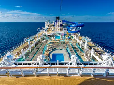 Blick von oben auf das Sonnendeck der Norwegian Bliss mit blauen Pools, vielen Liegestühlen und bunten Wasserrutschen. Im Hintergrund das weite Meer und der blaue Himmel.