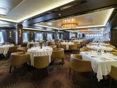 Weitläufiger, eleganter Speisesaal von Cagney's Steakhouse an Bord der Norwegian Bliss mit vielen gedeckten Tischen, beigen Lederstühlen und Blick aufs Meer durch große Fenster.