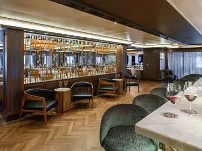 Die stilvolle Bar-Lounge des Spezialitätenrestaurants Cagney's an Bord der Norwegian Bliss. Moderne Ledersessel, ein Marmortresen mit Rotwein und eine dekorative Trennwand sind sichtbar.