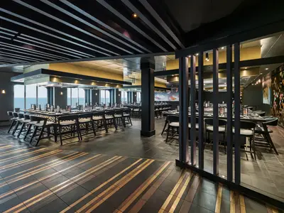 Modernes Teppanyaki-Restaurant an Bord der Norwegian Bliss. Lange Tische mit Stühlen und Meerblick. Dunkle, elegante Einrichtung mit goldenen Akzenten und Holzdetails.