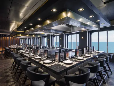 Das moderne Teppanyaki Restaurant an Bord der Norwegian Bliss. Elegant gedeckte Tische an Kochstationen. Grosse Fenster mit Meerblick. Dunkle Möbel.