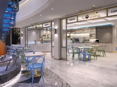 Das moderne Café „Coco’s“ an Bord der Norwegian Bliss. Helle Tische, bunte Stühle, Marmorboden, Süßspeisen-Theken. Links eine blau beleuchtete Wendeltreppe und Skulptur.