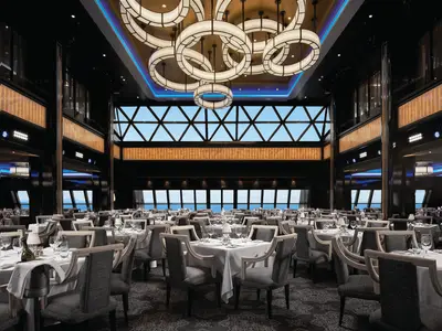 Blick in den eleganten Manhattan Room der Norwegian Bliss mit vielen weiß gedeckten Tischen und Stühlen. Große Fenster bieten einen weiten Blick auf das Meer und den Horizont. Stilvolle Deckenleuchten und Wandpaneele spenden warmes Licht. Der Raum ist menschenleer.