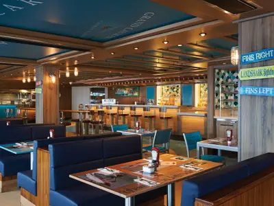 Innenansicht des Jimmy Buffett's Margaritaville Restaurants an Bord der Norwegian Bliss. Blaue Sitzbaenke, Holztische, Bar und maritime Deko sowie 'Fins Right' Wegweiser.