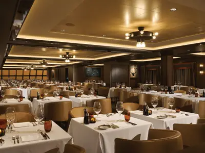 Elegantes Spezialitätenrestaurant Cagney's Steakhouse an Bord der Norwegian Bliss. Viele Tische mit weißen Tischdecken, Gläsern und braunen Ledersesseln. Warme Beleuchtung.