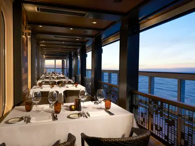 Elegantes Cagney’s Steakhouse an Bord der Norwegian Bliss mit Tischen am Fenster und Blick auf den ruhigen Ozean bei Sonnenuntergang.