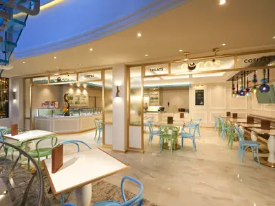 Modernes Coco's Dessert-Café an Bord der Norwegian Bliss. Helle Tische, bunte Stühle in Grün und Blau. Theken mit Schokoladen, Crepes und Kaffee im Hintergrund.