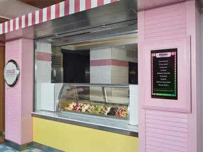 Detailansicht des Dolce Gelato Shops an Bord der Norwegian Bliss. Pink-gelbe Fassade mit rundem Logo. Eine Vitrine zeigt Gelato-Sorten; ein digitales Menü listet Geschmacksrichtungen.