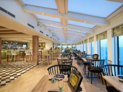 Modernes Food Republic Restaurant an Bord der Norwegian Bliss. Es zeigt helle Holzmoebel, schwarze Stuehle, Tische mit digitalen Menues, grosse Fensterfront mit Meerblick und offene Kueche.