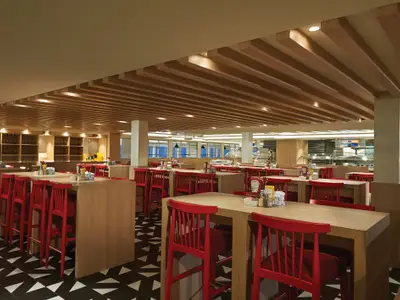Blick in das geräumige Garden Cafe Buffetrestaurant an Bord der Norwegian Bliss. Es zeigt Reihen von Holztischen mit roten Stühlen, dahinter Buffetauslagen und eine Weinbar. Die Decke ist mit Holzlamellen versehen.