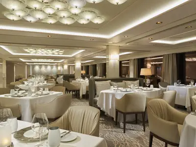 Elegantes, lichtdurchflutetes Spezialitätenrestaurant an Bord der Norwegian Bliss. Weiss gedeckte Tische mit Gläsern und Besteck, umgeben von hellen Sesseln. Moderne Beleuchtungselemente schmücken Decke und Wände.