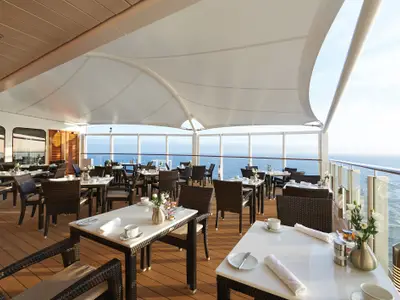 Blick auf den elegant gedeckten Aussenbereich des Restaurants The Haven an Bord der Norwegian Bliss mit zahlreichen Tischen und Stühlen unter einem Zeltdach. Im Hintergrund erstreckt sich das weite, blaue Meer bis zum Horizont.