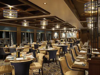 Elegantes Restaurant an Bord der Norwegian Bliss, abends. Gedeckte Tische, blaue und beige Stühle. Warme Deckenleuchten und große Fenster schaffen eine einladende Atmosphäre.