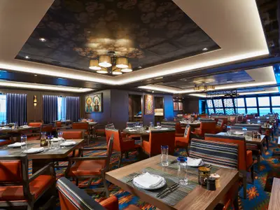 Blick in das farbenfroh gestaltete Restaurant Los Lobos an Bord der Norwegian Bliss. Zahlreiche Tische und Stühle in Erdtönen, kunstvolle Decken und große Fenster mit Meerblick sind zu sehen.