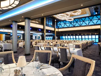 Geräumiger, elegant gedeckter Speisesaal im Manhattan Room der Norwegian Bliss mit vielen Tischen und Stühlen. Große Fenster bieten einen weiten Blick auf das Meer. Moderne Beleuchtung und Säulen mit Mosaikverkleidung prägen das stilvolle Ambiente.