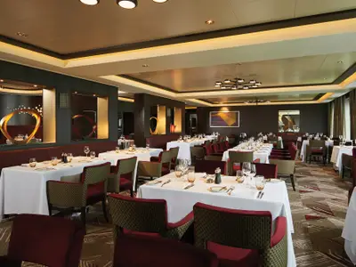Ein stilvolles Restaurant an Bord der Norwegian Bliss mit zahlreichen gedeckten Tischen, roten Stühlen und gemütlicher Beleuchtung. Moderne Kunstobjekte schmücken die Wände, und warme Holzelemente prägen das Ambiente.