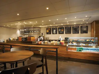 Innenansicht des Starbucks auf der Norwegian Bliss mit Theke, Kaffee- und Snackauslage, Getränkekühlschrank, Menütafeln und Holzmöbeln.