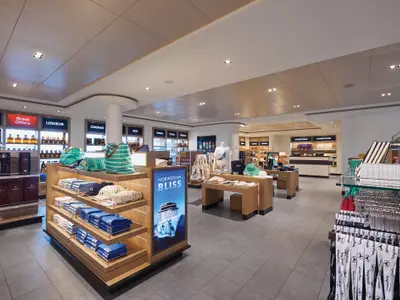 Moderner Duty-Free-Shop Tradewinds an Bord der Norwegian Bliss. Regale mit Spirituosen und Souvenirs wie Kleidung und Taschen. Ein Display wirbt für das Schiff.