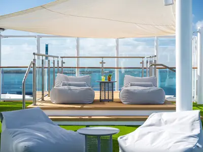 Ein sonniger Entspannungsbereich auf dem Deck der Norwegian Bliss mit grauen Sitzsäcken und einem Tisch mit zwei Cocktails. Im Hintergrund ist das Meer sichtbar.