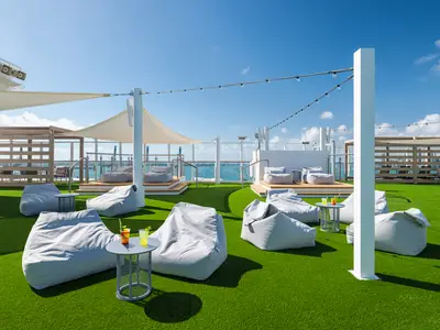 Blick auf das Horizon Park Sonnendeck der Norwegian Bliss. Graue Bean Bags auf grünem Kunstrasen mit Tischen, Sonnensegeln und Meerblick.