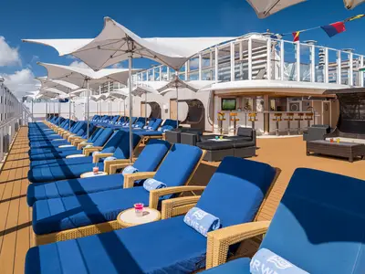 Sonniges Deck der Norwegian Bliss mit blauen Liegestühlen, weissen Sonnenschirmen und einer Bar. Blauer Himmel, entspannte Atmosphäre an Bord.