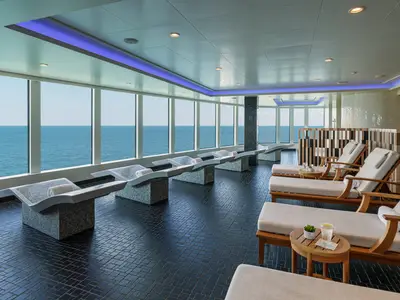 Der luxuriöse Wellnessbereich an Bord der Norwegian Bliss bietet komfortable Thermal- und Holz-Lounger mit Meerblick durch große Fenster. Entspannte Atmosphäre mit stilvoller Einrichtung und blauer Lichtinstallation.