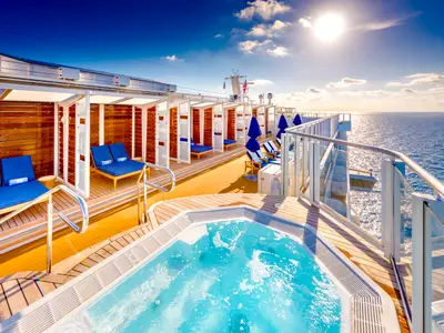 Ein Whirlpool und blaue Sonnenliegen in modernen Holz-Cabanas auf dem Sonnendeck der Norwegian Bliss. Im Hintergrund glitzert das Meer bei strahlendem Sonnenschein.