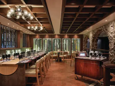 Elegante Weinkeller-Bar an Bord der Norwegian Bliss. Langer Tisch für Weinproben mit Gläsern und Snacks. Weinschränke und rustikale Steinwand im Hintergrund. Warme Beleuchtung.