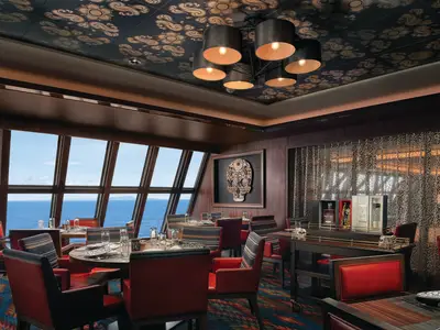 Elegantes Los Lobos Spezialitätenrestaurant an Bord der Norwegian Bliss mit Meerblick. Der Innenbereich zeigt stilvolle Möbel, gedeckte Tische und eine auffällige Totenkopf-Skulptur an der Wand.