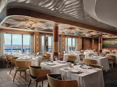 Innenansicht des eleganten Spezialitätenrestaurants Ocean Blue an Bord der Norwegian Bliss. Zahlreiche Tische mit weissen Tischdecken und Blick auf das Meer durch grosse Fenster sind zu sehen.