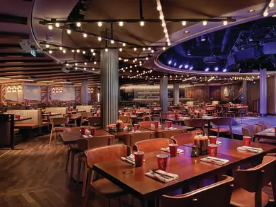 Das Q Texas Smokehouse Restaurant an Bord der Norwegian Bliss. Viele Tische, rote Becher, Ledersitze und eine Bühne für Live-Musik sind zu sehen. Lichterketten und Western-Wandgemälde dominieren den Raum.