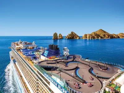 Vogelperspektive auf die Norwegian Bliss, die auf dem offenen Meer vor felsigen Inseln in Mexiko fährt. Auf dem Oberdeck befinden sich eine Go-Kart-Rennstrecke und blaue Wasserrutschen.
