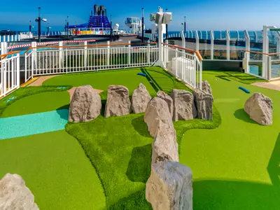 Mini-Golfanlage an Deck der Norwegian Bliss: grüner Kunstrasen, Felsen, blaues Wasserhindernis. Im Hintergrund Meer und Schiffsaufbauten.