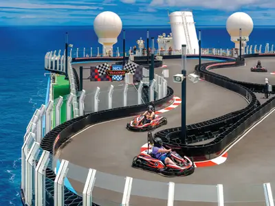 Vogelperspektive auf die Go-Kart-Rennstrecke auf dem Oberdeck der Norwegian Bliss. Mehrere Personen fahren rote Karts auf der kurvigen Strecke. Im Hintergrund ist das weite blaue Meer zu sehen.